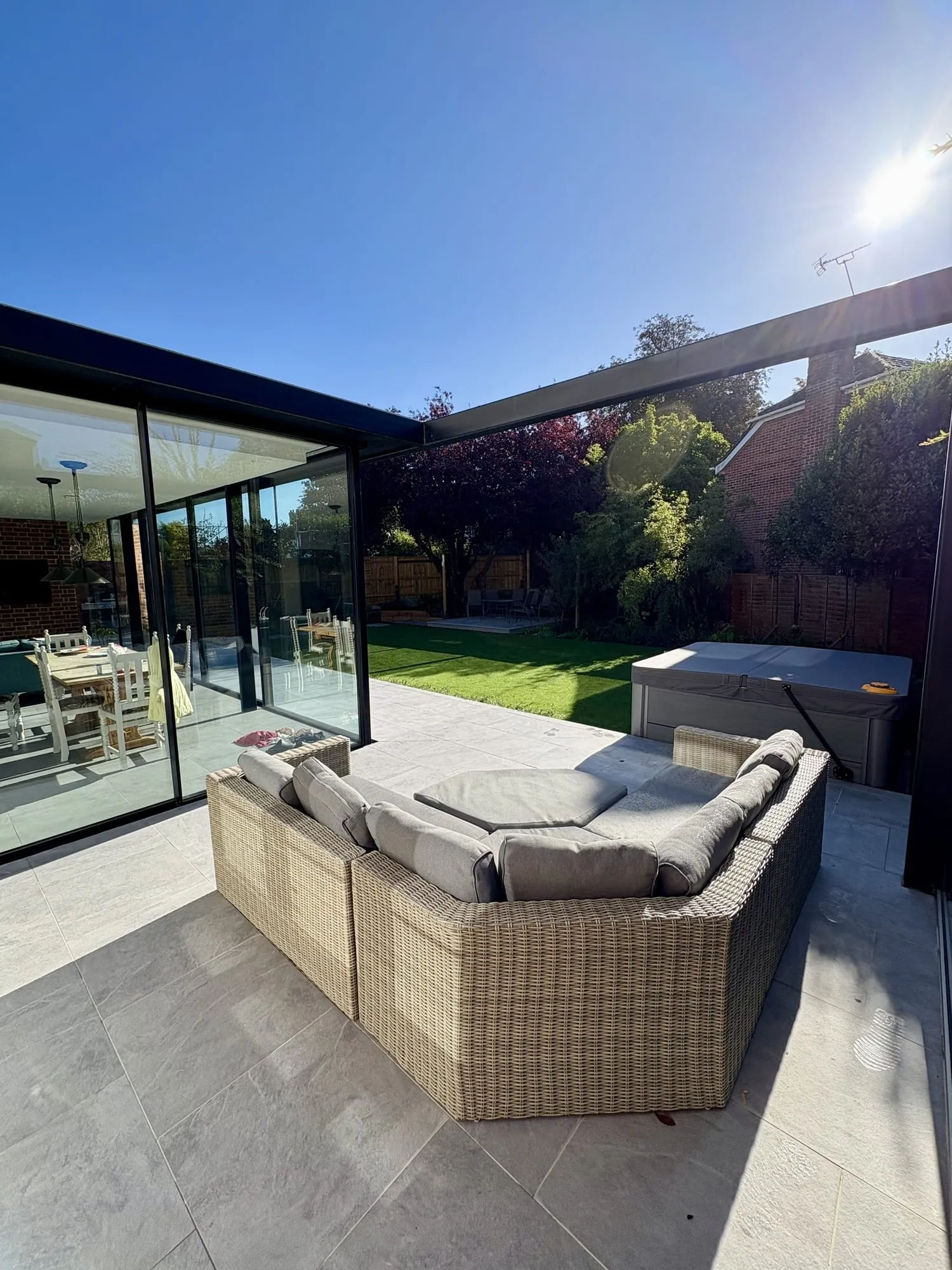 Rattan corner sofa on a porcelain patio beneath a pergola, sun flaring through the frame