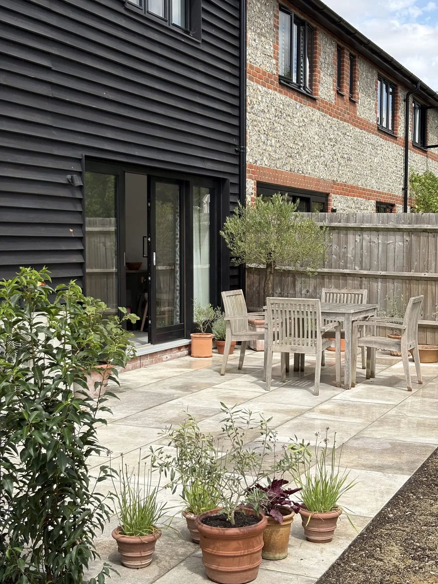 Timber table and chairs on a paved terrace beside a black clad house, with potted planting